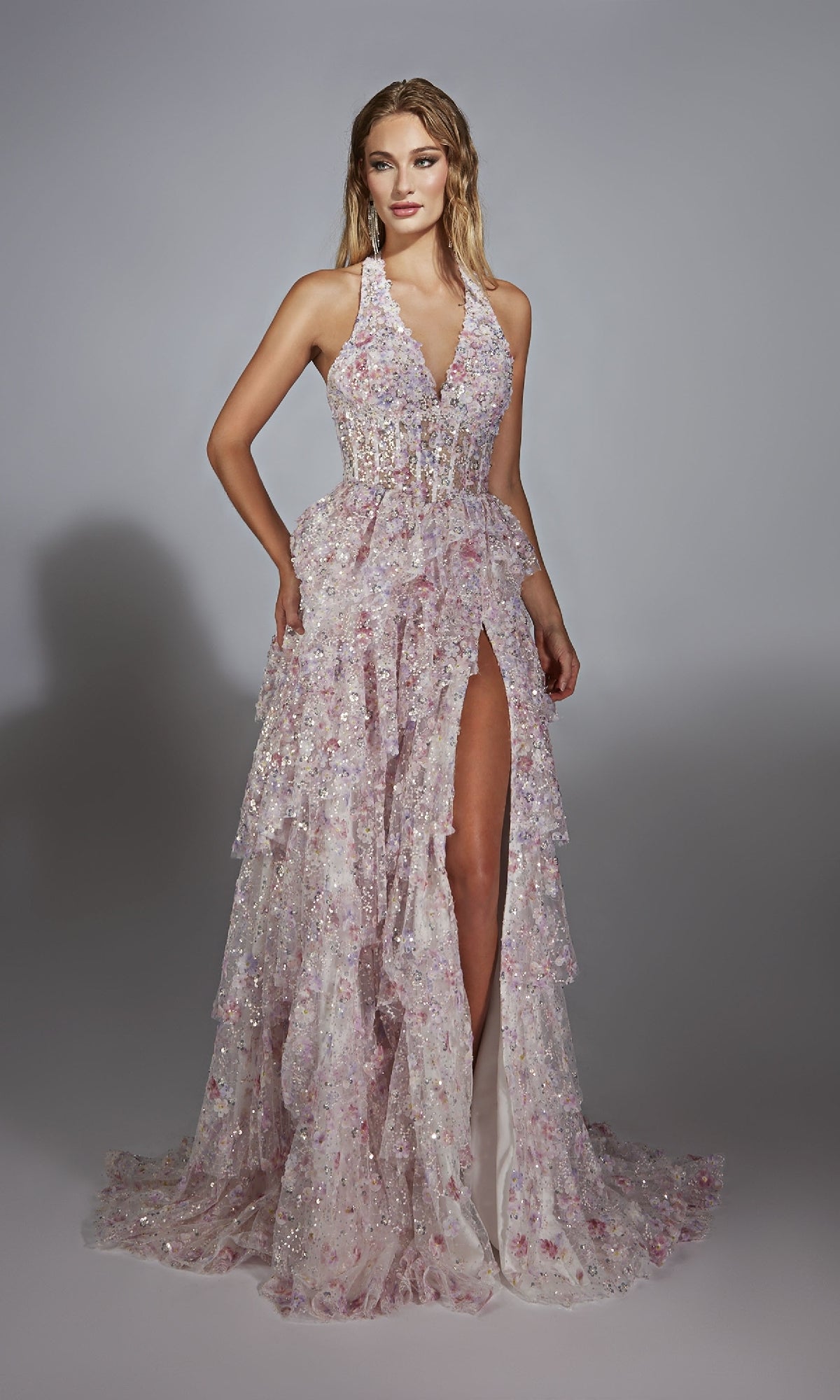 A woman models the Alyce 61740 long prom dress—a light pink gown with a plunging neckline, high slit, ruffled layers, and sparkling floral embellishments—set against a neutral background.